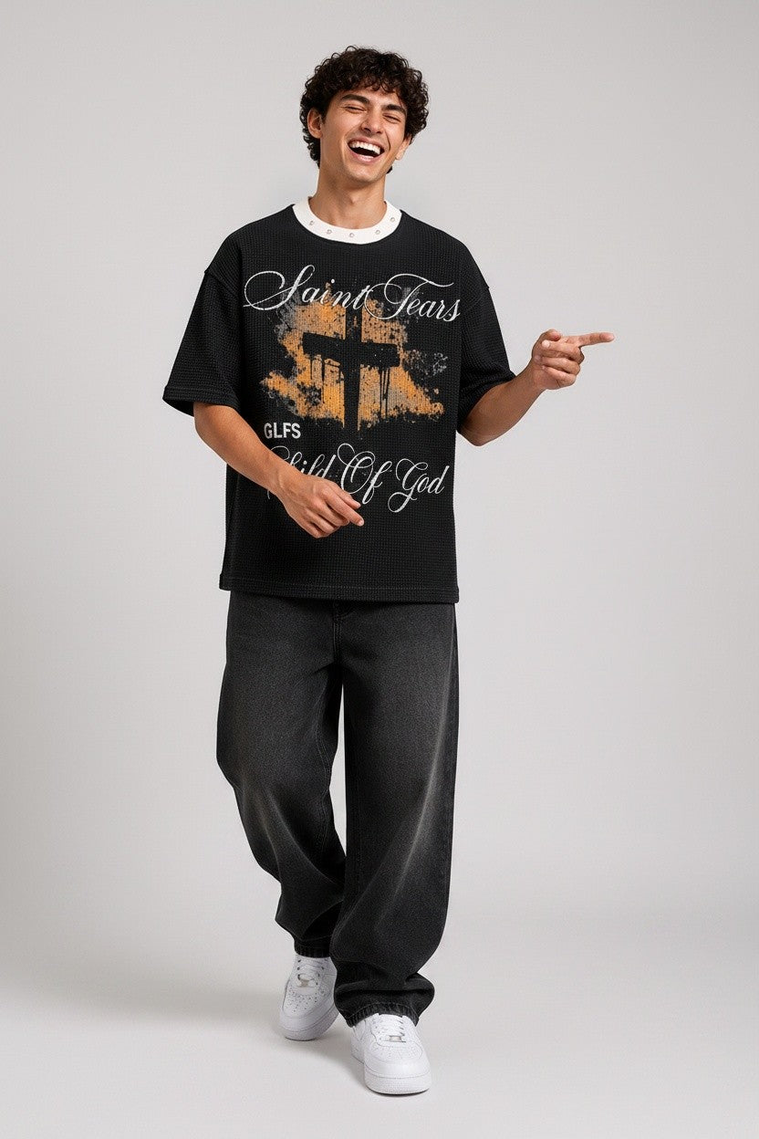 PAKSHI black waffle knit t-shirt with a graphic cross print, worn by a smiling man in loose-fit charcoal jeans.
